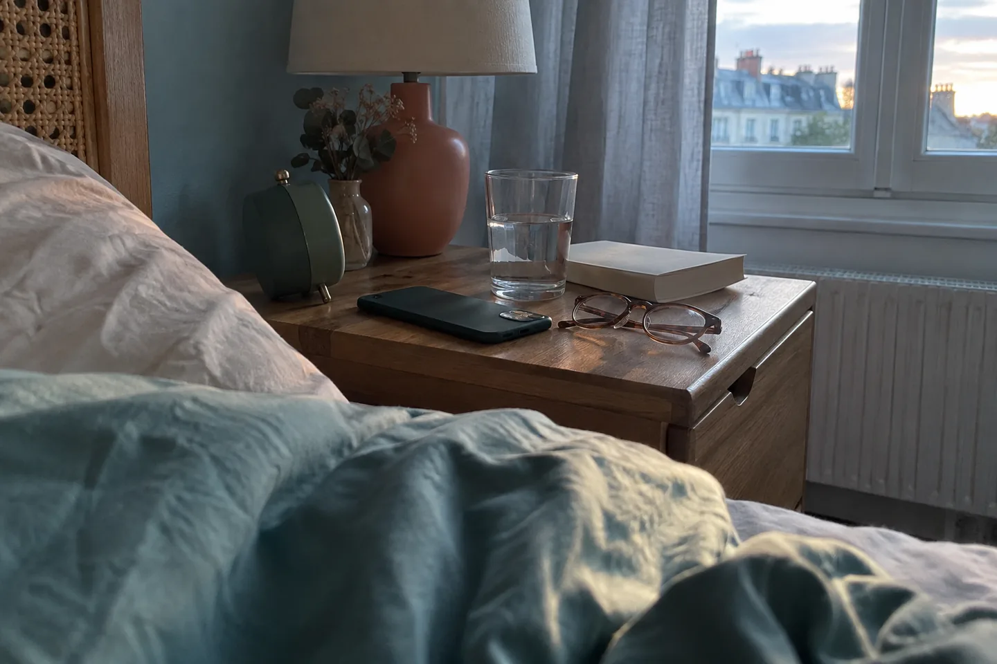 Sur une table de nuit, un téléphone posé écran contre le bois, à côté d’un verre d’eau, d’un livre fermé et d’un petit réveil.