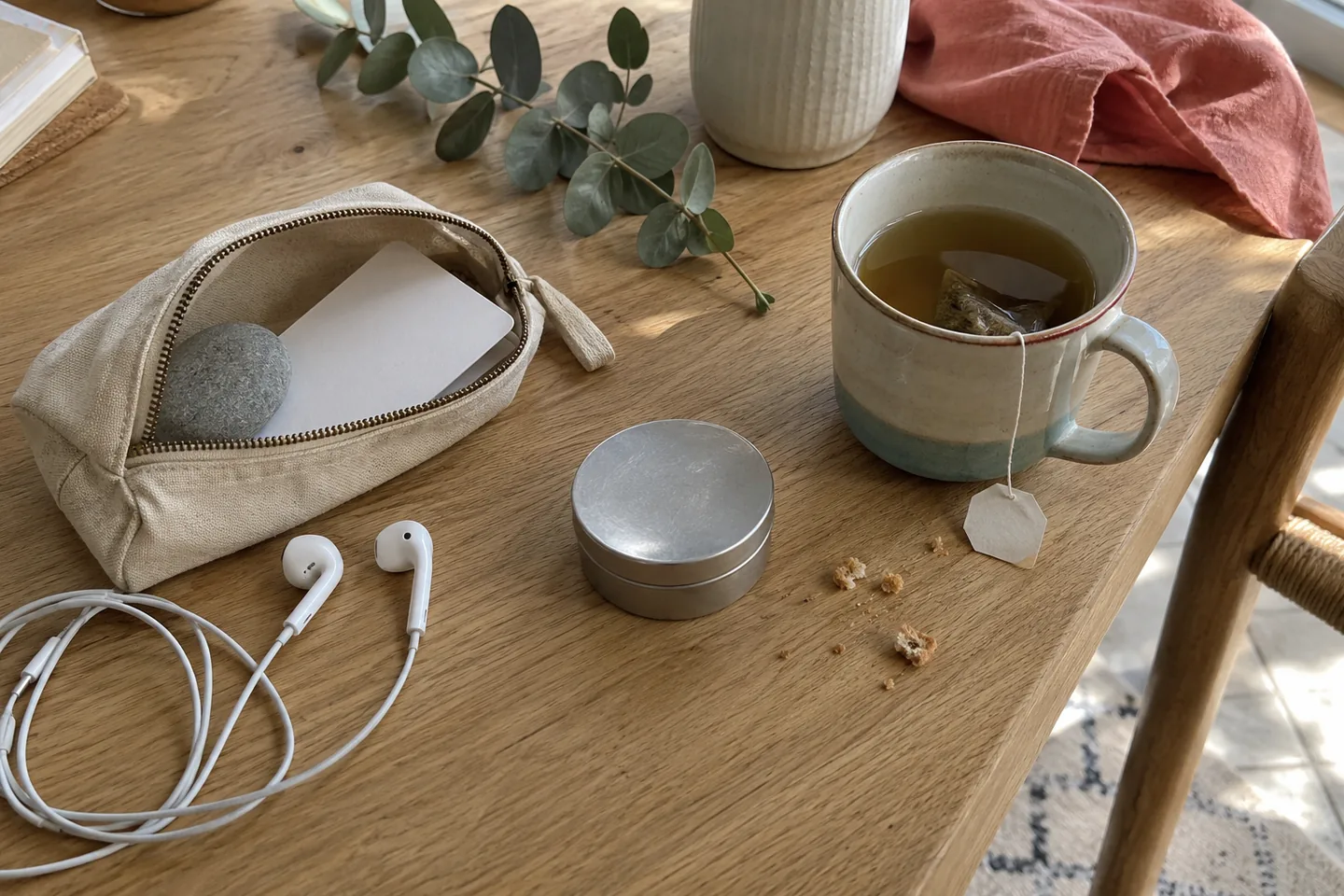 Une petite trousse en tissu ouverte sur une table, avec un galet lisse, une carte vierge pliée, des écouteurs et une tasse de tisane.